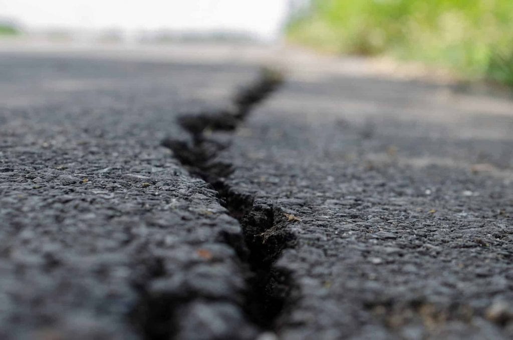 Close-up of a large crack running through the surface of an asphalt road in NJ, with blurred greenery on the right side—expertly documented by Paving Contractors Morris serving Sussex & Somerset County.