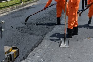 Workers in orange uniforms and black boots are busy with residential paving, spreading asphalt on a road with shovels. The section being resurfaced is partially completed, set against the backdrop of a sunny day.