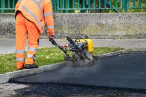 A worker in orange safety gear operates a vibrating plate compactor on fresh asphalt, a hallmark of professional residential paving. Dust and steam rise from the newly laid pavement, with a concrete barrier and green fence visible in the background.