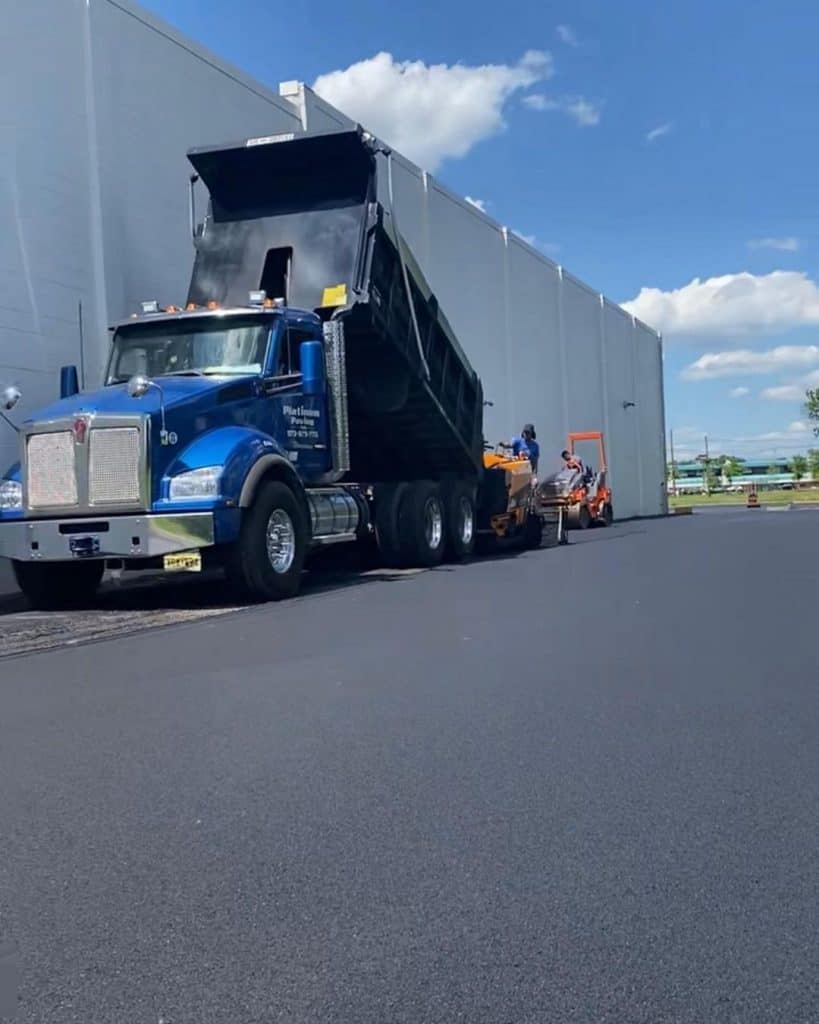A blue dump truck is unloading black asphalt onto a freshly paved surface next to a large white building. Paving contractors expertly operate machinery to spread and smooth the asphalt under a partly cloudy sky.