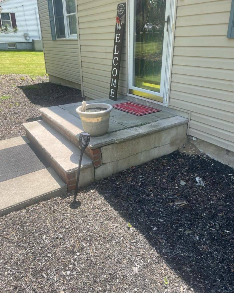 A small front porch with two expertly crafted concrete steps leading to a glass door. A vertical wooden welcome sign leans against the house. A pot with water and a solar garden light stand on the porch designed by skilled paving contractors. The ground is covered with mulch.