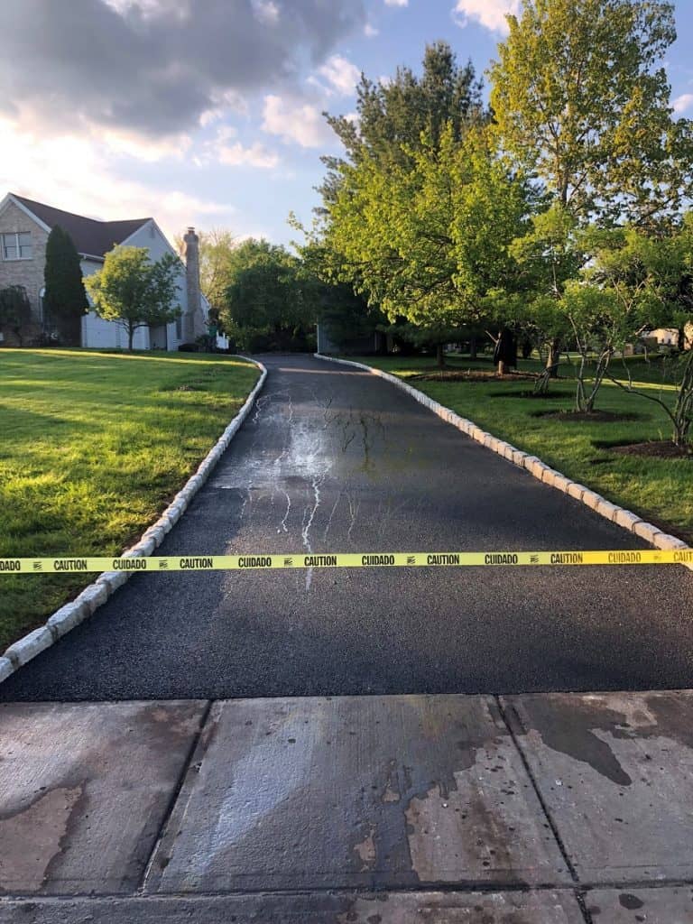 A newly paved driveway is blocked by yellow caution tape, with wet surface marks visible. Green trees and grass surround the area, and a house is seen in the background under a partly cloudy sky.
