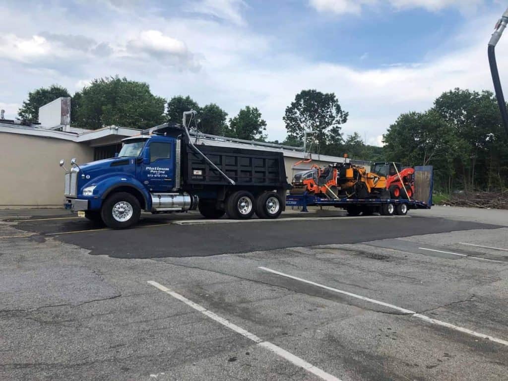 A blue dump truck with a black bed, used by Paving Contractors Morris, NJ, is parked in a lot towing a flatbed trailer with orange construction equipment. Trees and a building are visible in the Sussex & Somerset County background.