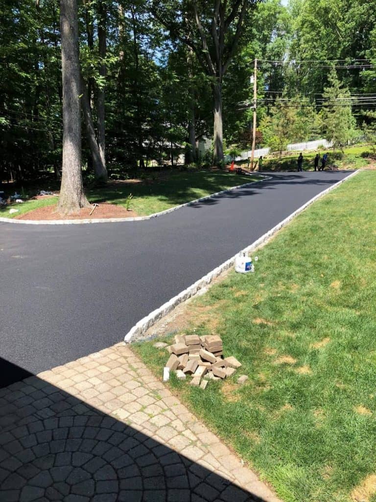 Freshly paved black asphalt driveway curves through a green lawn, bordered by stone edging. Paving Contractors Morris, Sussex & Somerset County, NJ have tools and paving stones visible on the grass, all surrounded by tall trees and a sunny sky.