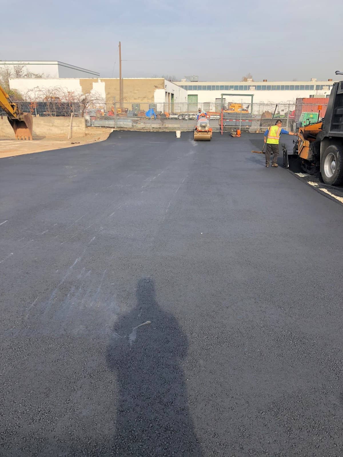 Freshly paved asphalt road with a construction worker operating a roller in the distance, another worker near a truck on the right, and construction equipment and barriers in the background. The photographer’s shadow is visible.