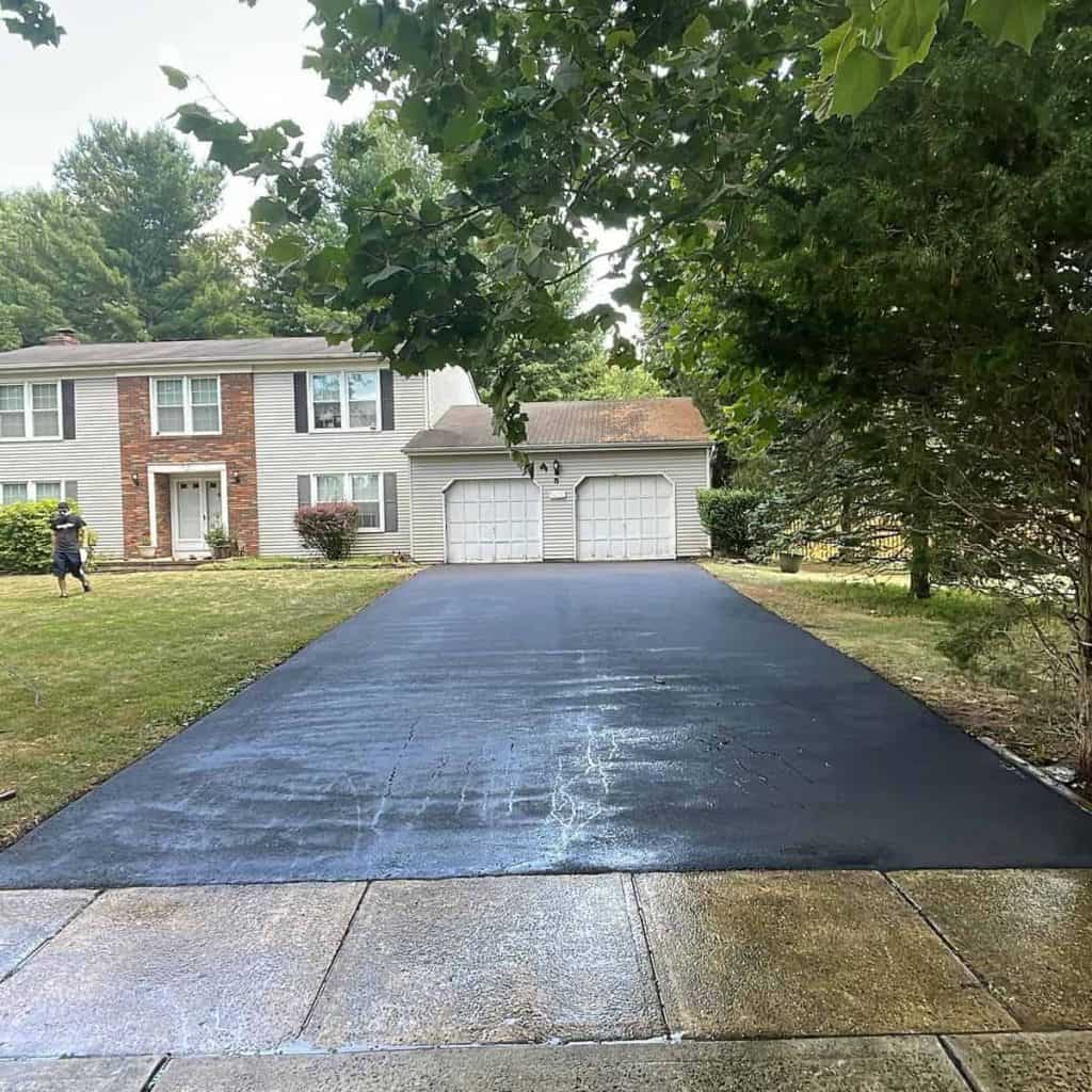 A freshly paved black asphalt driveway by Paving Contractors Morris, NJ, leads to a two-car garage attached to a light-colored suburban house with brick accents, surrounded by grass, trees, and wet pavement in the foreground.