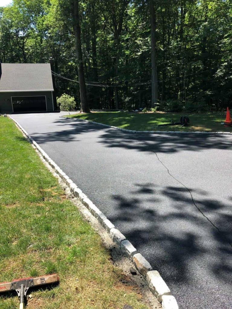 A newly paved asphalt driveway by Paving Contractors Morris leads to a garage, bordered by grass and a stone curb, with trees casting shadows on the surface—serving homes across Sussex & Somerset County, NJ.