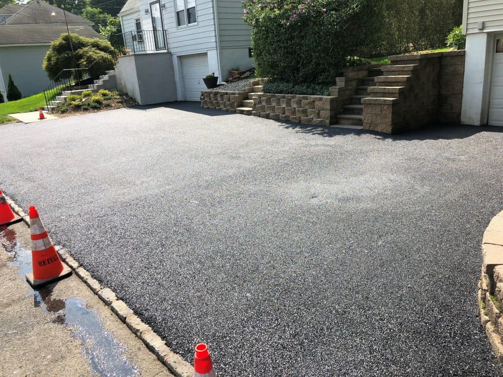 Freshly paved black asphalt driveway in front of a house with a concrete staircase, stone retaining wall, and orange traffic cones at the edges to block entry.