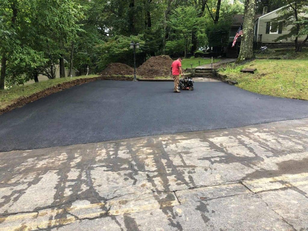 A person in a red shirt operates machinery on a newly paved asphalt driveway, showcasing the expertise of Paving Contractors Morris, NJ, with dirt piles and trees in the background near a house.