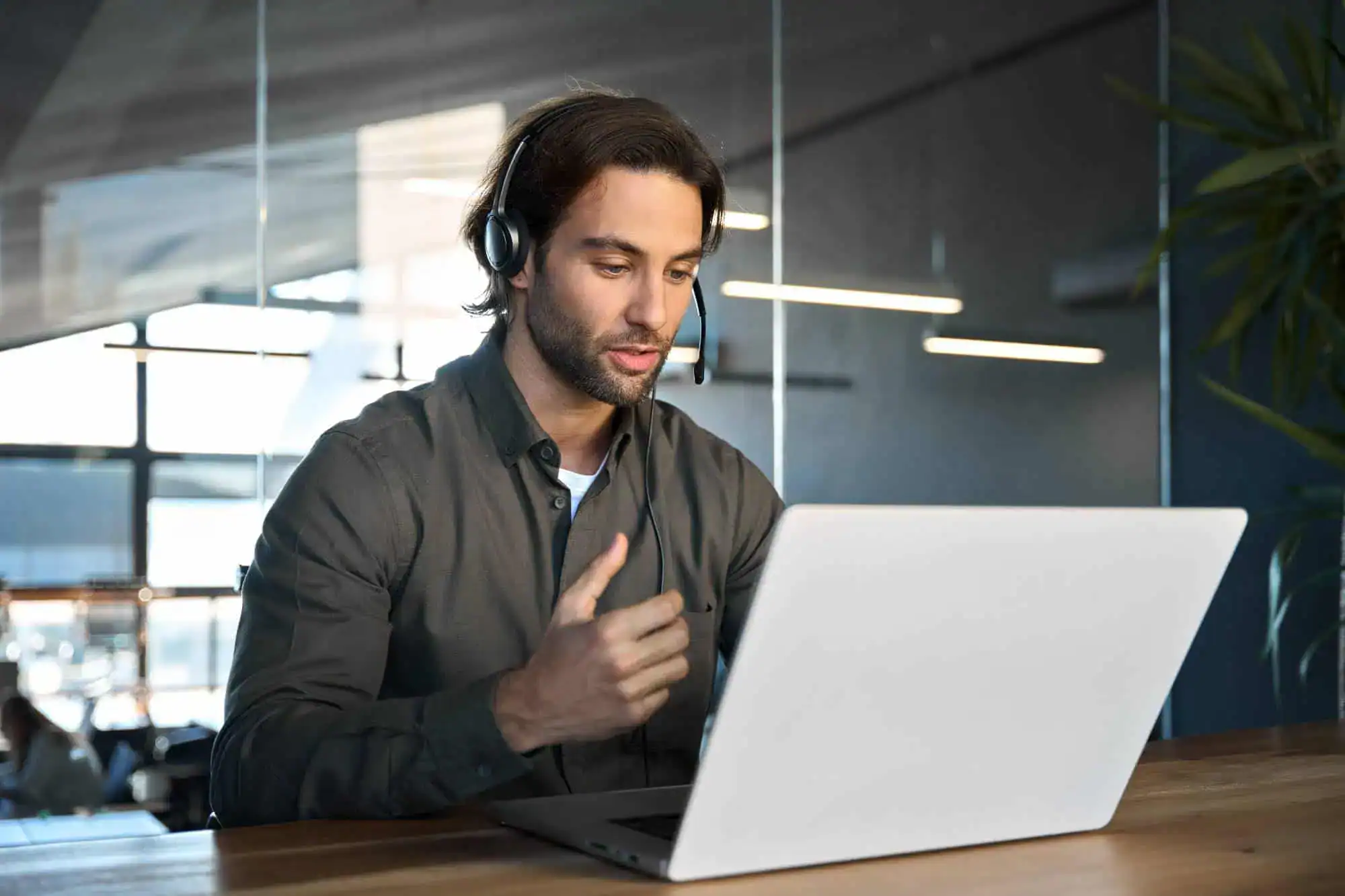 A man wearing a headset sits at a desk in front of a laptop, gesturing with his hand while participating in a video call with Paving Contractors Morris, NJ, in a modern Sussex & Somerset County office setting.