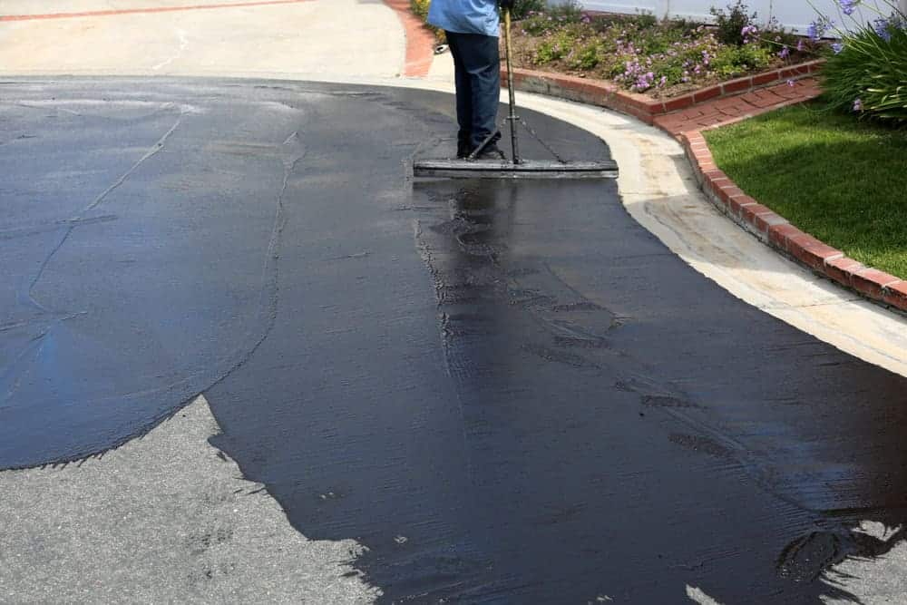 A person uses a long-handled tool to apply black sealcoat to a curved driveway in NJ, with freshly coated and uncoated areas visible—quality work from paving contractors in Morris.