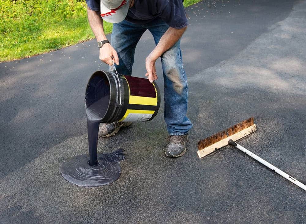 A person pours black sealant from a large bucket onto an asphalt driveway, preparing to spread it—just like the experts at Paving Contractors Morris, Sussex & Somerset County, NJ. A broom for spreading sealant lies on the ground nearby.