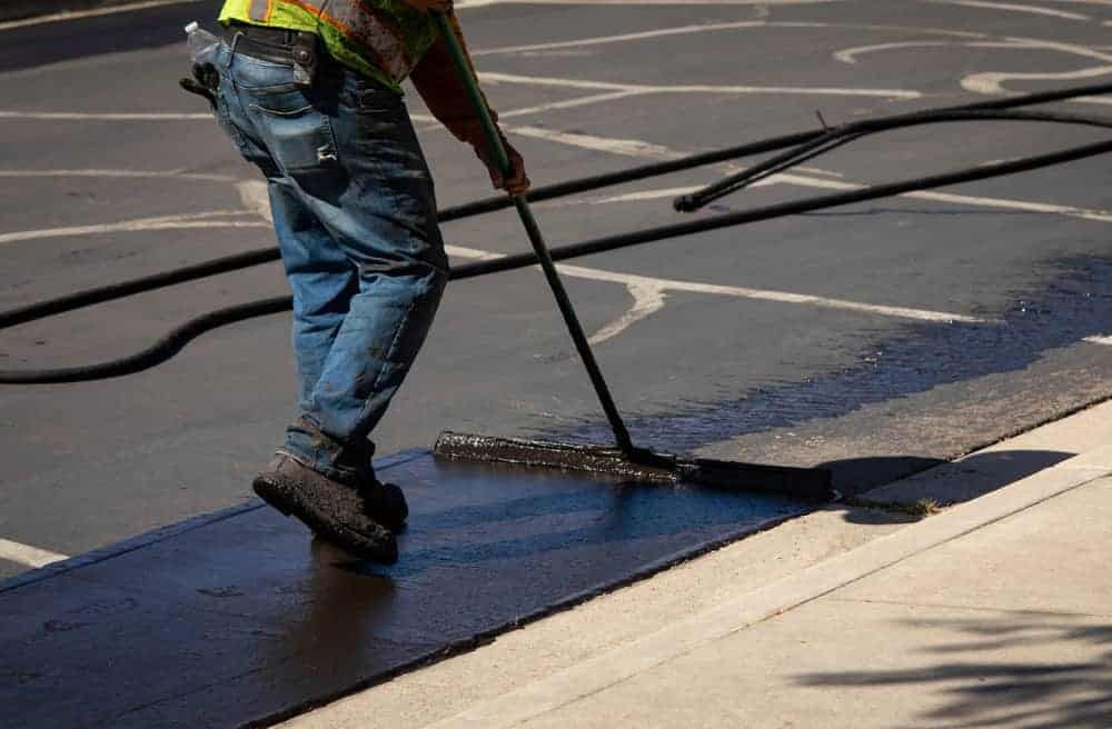 A worker in a safety vest and jeans uses a large squeegee to apply black sealant to an asphalt parking lot, with marked parking spaces visible—quality work by paving contractors serving Morris, Sussex & Somerset County, NJ.