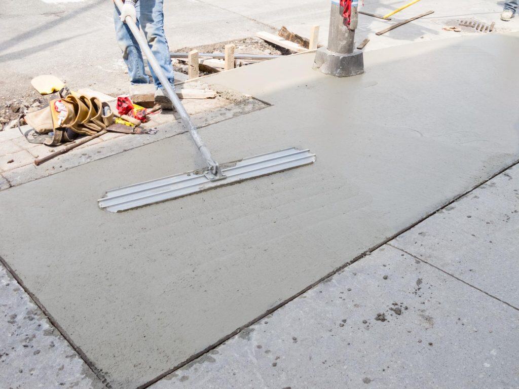 A worker uses a long-handled tool to smooth freshly poured concrete on a sidewalk. Paving Contractors Morris, NJ, Sussex & Somerset County ensure quality work, with construction materials, safety vest, and gloves visible nearby.