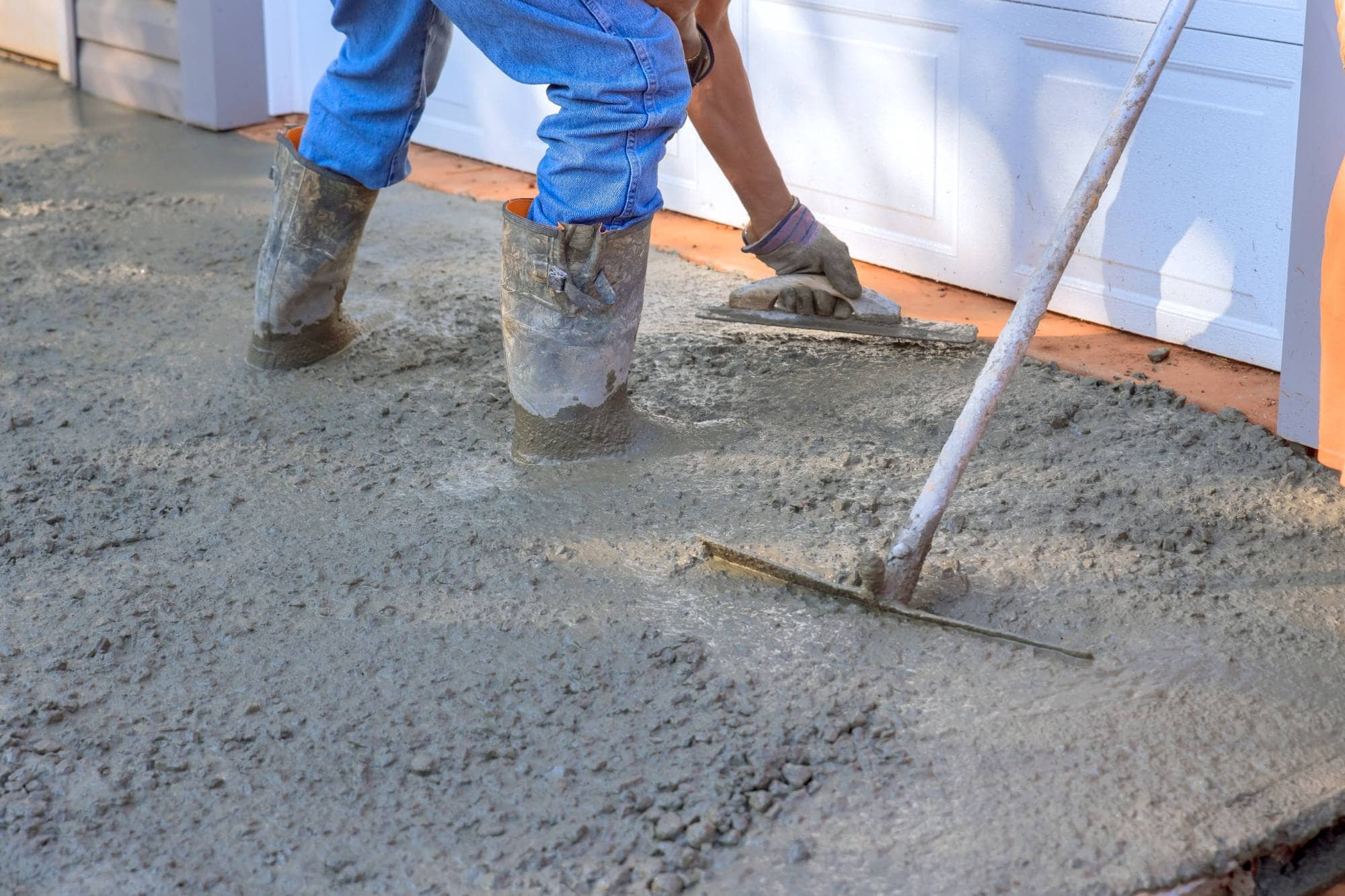 A person wearing muddy boots and blue jeans levels wet concrete with a trowel and a rake in front of a white garage door—typical of paving contractors in Sussex & Somerset County, NJ.