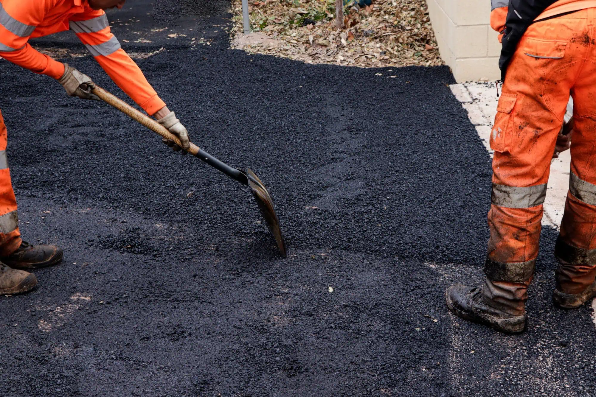 Two workers in orange safety gear spread and level freshly laid asphalt on a road with shovels. Only their lower bodies are visible, and the asphalt appears dark and newly applied.