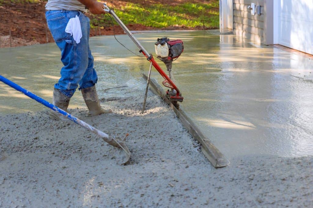 A worker in jeans and boots smooths wet concrete with a power trowel while another tool levels the surface, as Paving Contractors Morris prep a driveway or patio near a house in sunny Sussex & Somerset County, NJ.