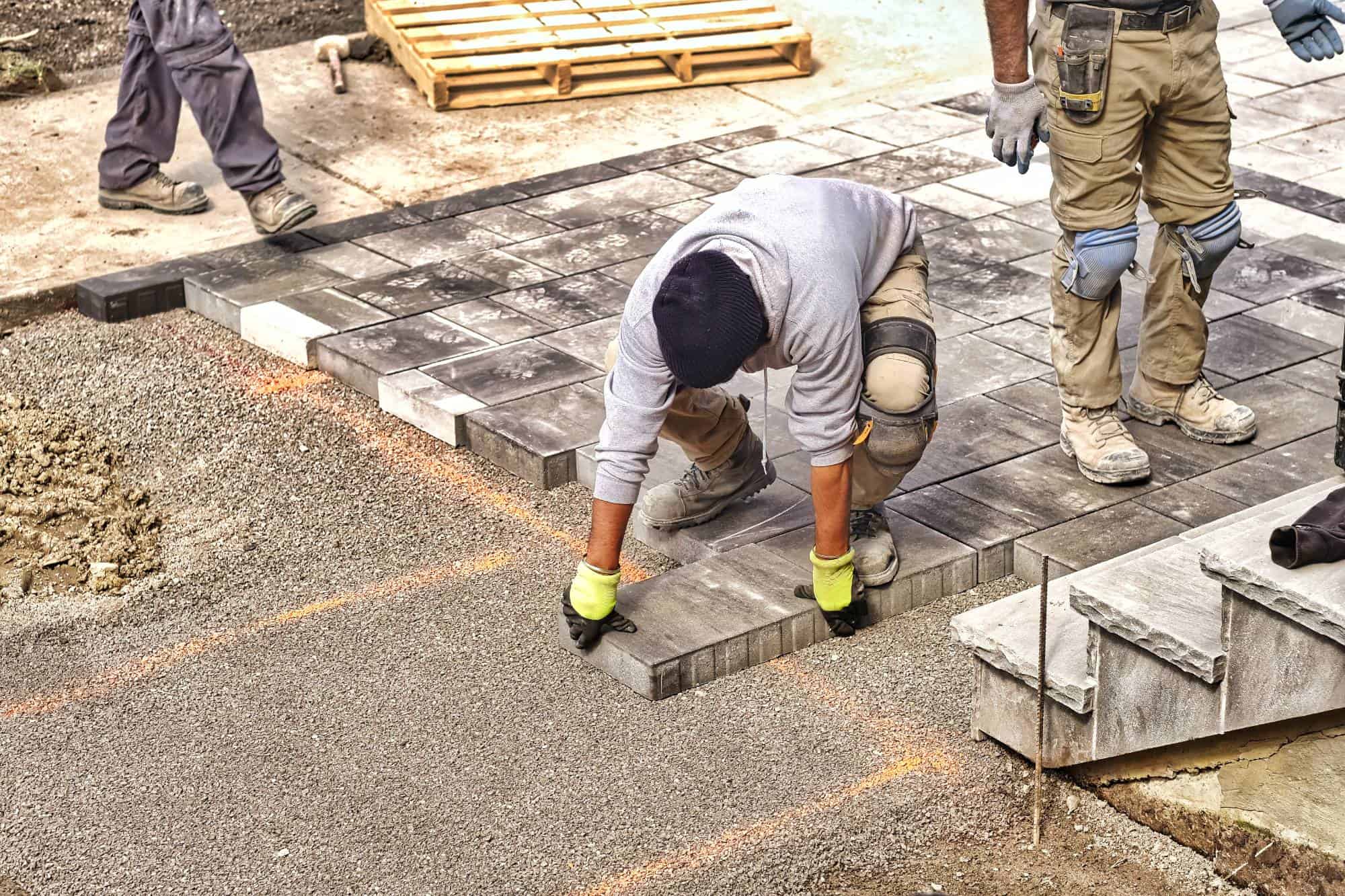 A worker in gloves and kneepads installs rectangular paving stones on a gravel surface while another stands nearby. Construction materials and tools are visible—expert paving contractors serving Morris, Sussex & Somerset County, NJ.