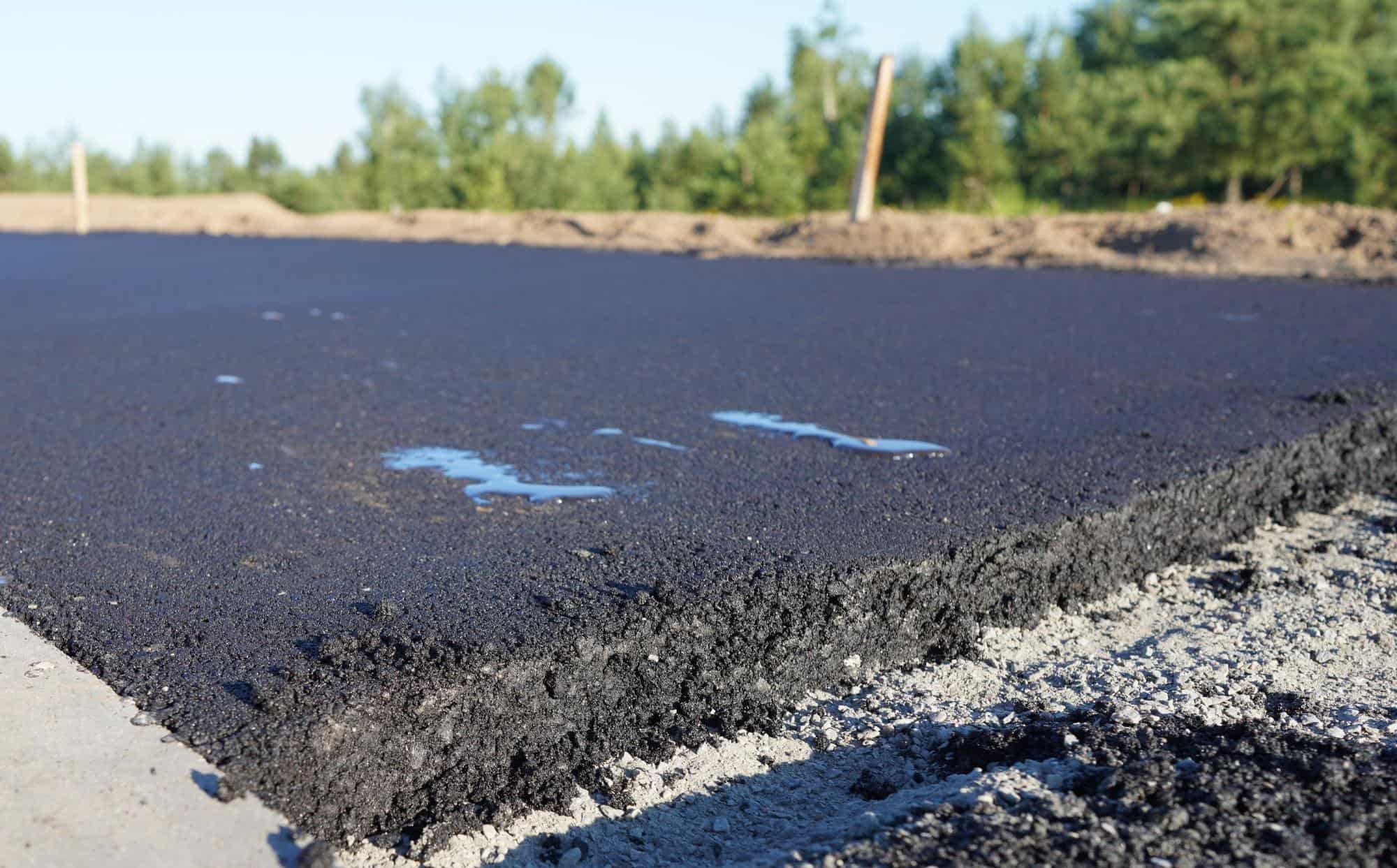 Freshly laid asphalt pavement by paving contractors in Morris, Sussex & Somerset County, NJ, with a close-up view of its rough, black surface. Small puddles of water rest on top as trees and soil blur in the background.