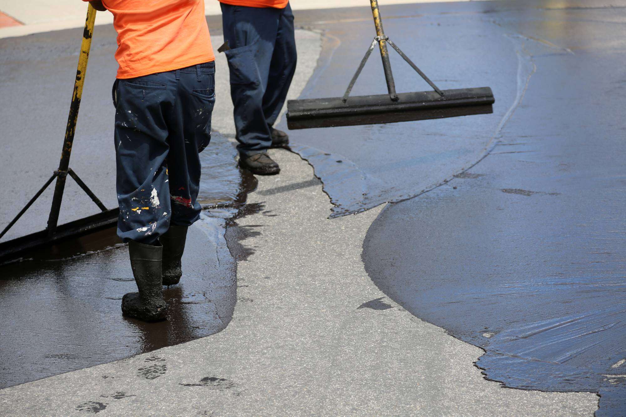 Two workers in orange shirts and boots use large squeegees to spread fresh asphalt on a road in Sussex & Somerset County, NJ, covering the grey surface with a smooth, dark layer. Only their lower bodies are visible&mdash;true Paving Contractors Morris professionals at work.