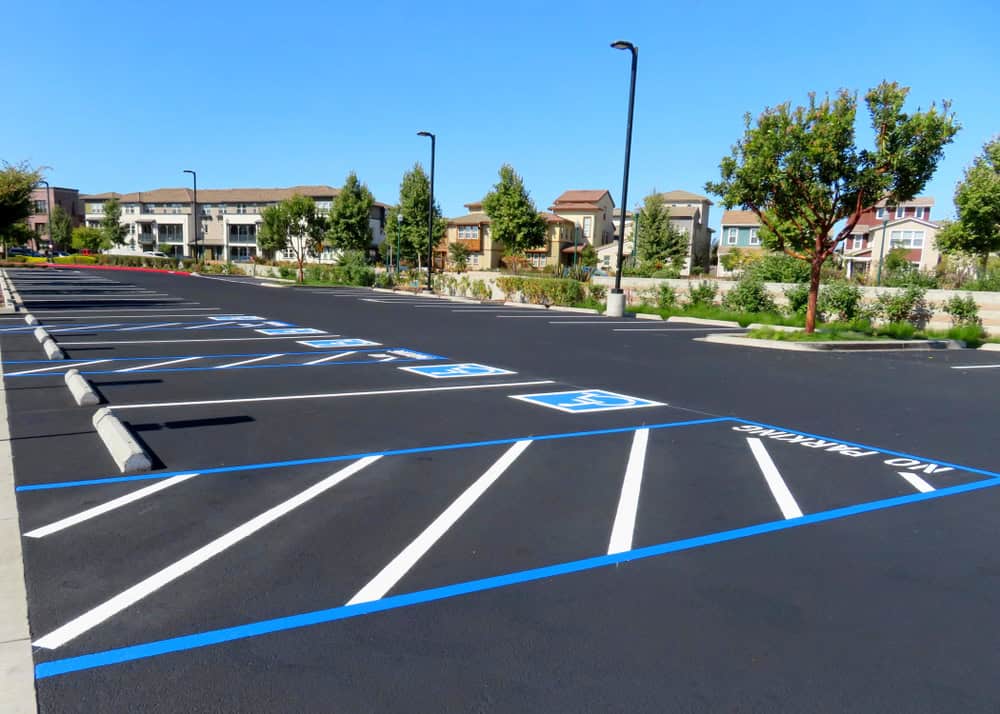 Accessible parking spaces with blue markings and "NO PARKING" zones are in an empty parking lot near a landscaped area and residential buildings under a clear blue sky.