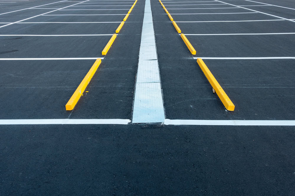 Empty parking lot with black asphalt, white painted lines marking parking spaces, yellow wheel stops, and a metal drainage grate running down the center.
