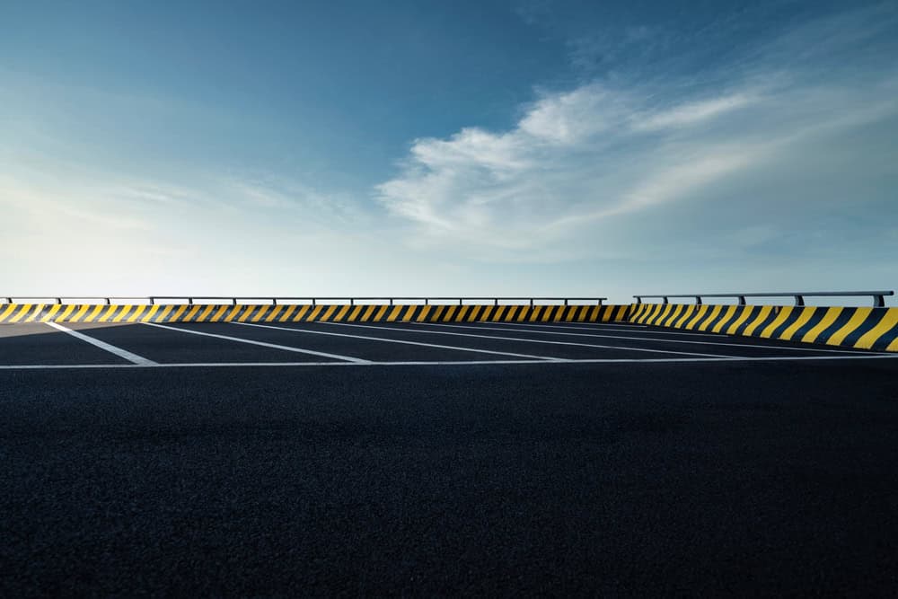 Empty parking lot with white lines on dark asphalt, bordered by a low concrete barrier with black and yellow diagonal stripes, under a blue sky—expertly finished by Paving Contractors Morris, NJ, Sussex & Somerset County.