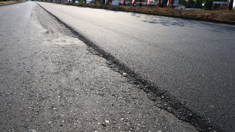 A close-up view of a road under construction, showing a clear divide between the new smooth asphalt on the left and the rough, older surface on the right, with buildings visible in the background.