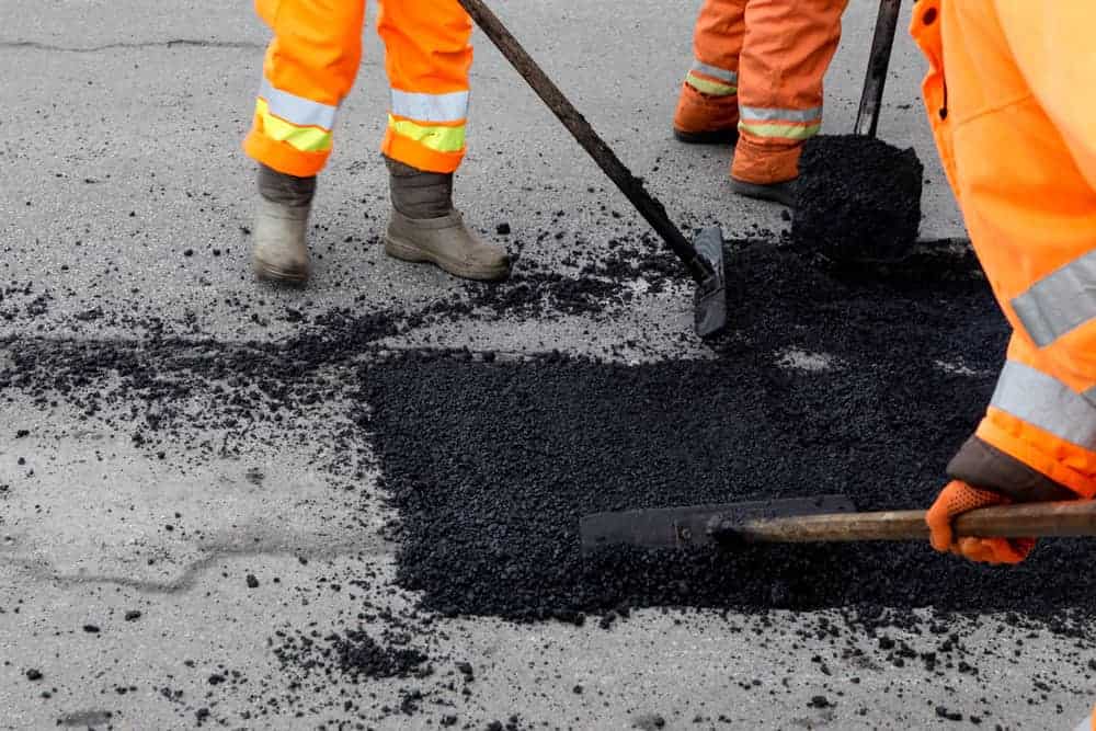 Workers in orange reflective clothing from Paving Contractors Morris use tools to spread and smooth black asphalt on a road surface in Sussex & Somerset County, NJ. Only their lower bodies and tools are visible in the image.