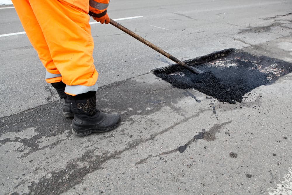 A road worker in orange pants and black boots uses a tool to spread fresh asphalt into a pothole on a cracked street, repairing the road surface.