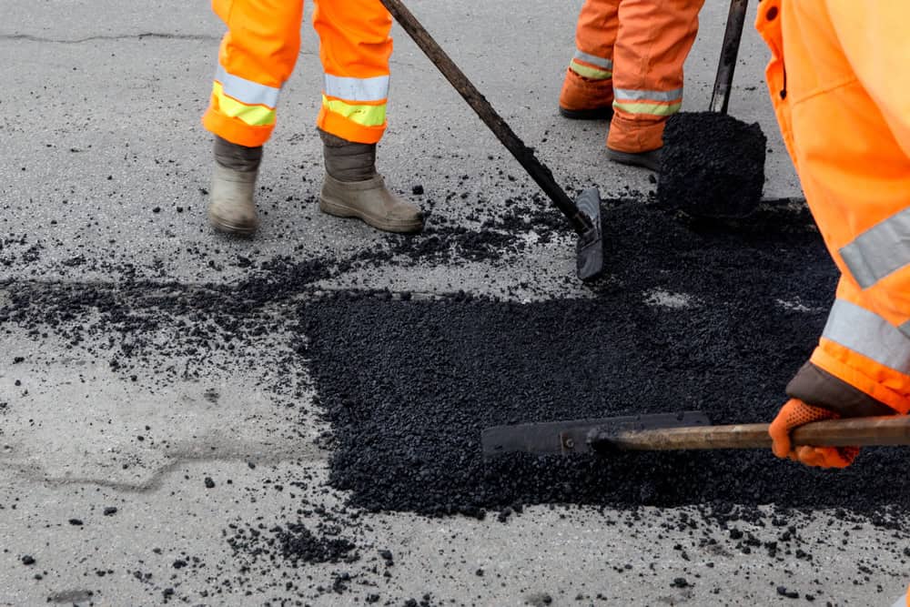 Workers in orange reflective clothing use shovels to spread asphalt while repairing a road. The image focuses on their legs and tools as they patch a section of damaged pavement.