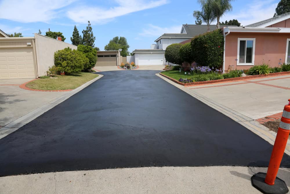 A freshly paved asphalt driveway runs between two suburban homes, leading to white garage doors. Flowerbeds and green shrubs border the driveway, and an orange safety cone sits at the entrance. The sky is partly cloudy.