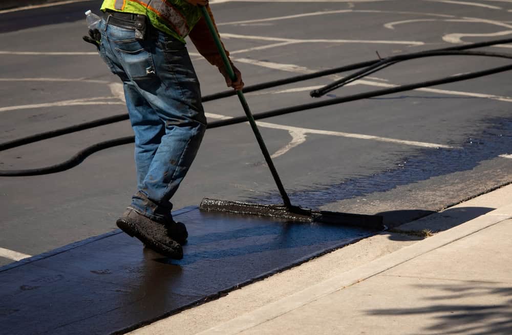 A worker in jeans and a safety vest uses a long-handled tool to apply sealant to an asphalt parking lot, resurfacing the pavement near a curb.