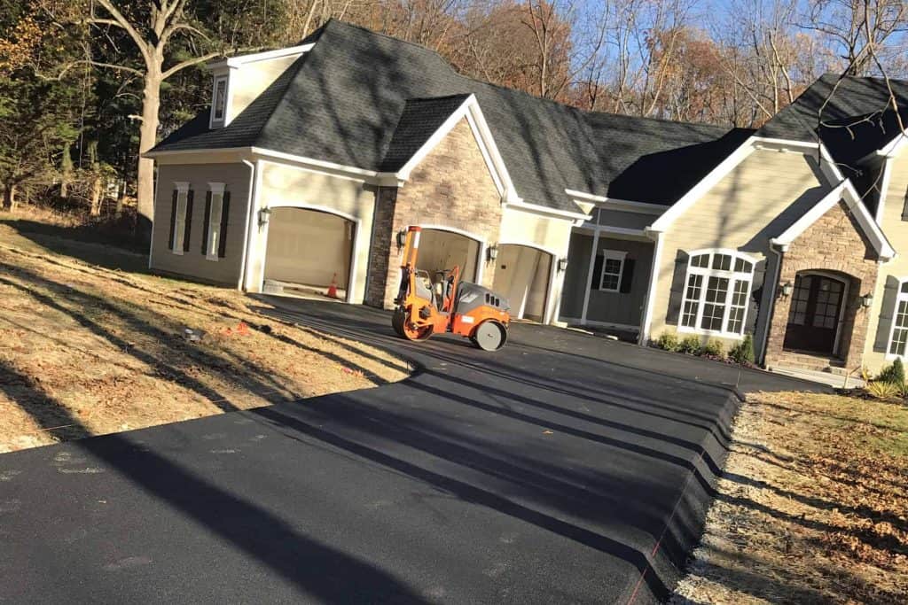 A new asphalt driveway in NJ is being compacted by an orange roller in front of a large, two-story house with light siding and a two-car garage, surrounded by trees. Paving Contractors Morris serve Sussex & Somerset County.
