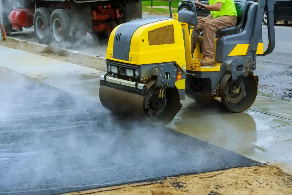 A worker operates a yellow road roller to flatten and smooth freshly laid asphalt on an NJ road, with steam rising from the hot surface. A truck and green grass are visible, showcasing Paving Contractors Morris, Sussex & Somerset County at work.