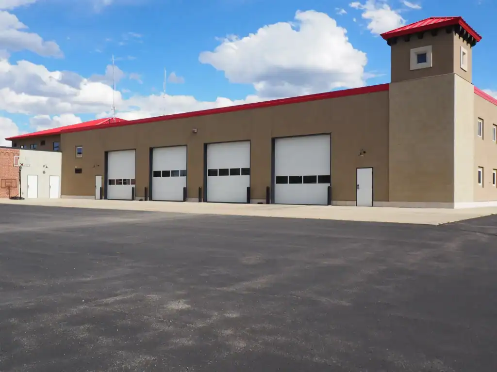 A modern fire station with four large garage doors and a small tower on the right, featuring tan walls and a red roof, stands under a blue sky in Sussex & Somerset County, NJ—expertly finished by Paving Contractors Morris.