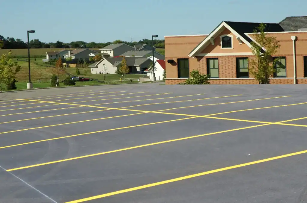 An empty parking lot with freshly painted yellow lines stretches before a brick building, expertly finished by paving contractors in Morris, Sussex & Somerset County, NJ. In the background are houses, green lawns, trees, and a clear blue sky.