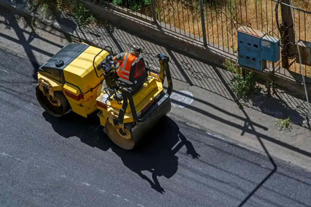 A construction worker in an orange safety vest operates a yellow road roller, compacting fresh asphalt along a street—expert work by paving contractors from Morris, NJ, Sussex & Somerset County—next to a fence and utility box on a sunny day.