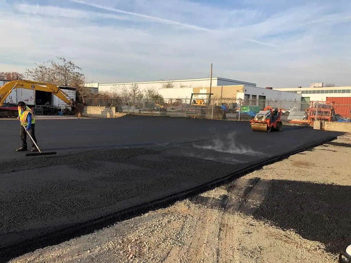A construction worker spreads asphalt with a rake while another operates a steamroller on a large outdoor site, as Paving Contractors Morris, NJ lay new pavement near industrial buildings under a partly cloudy sky.