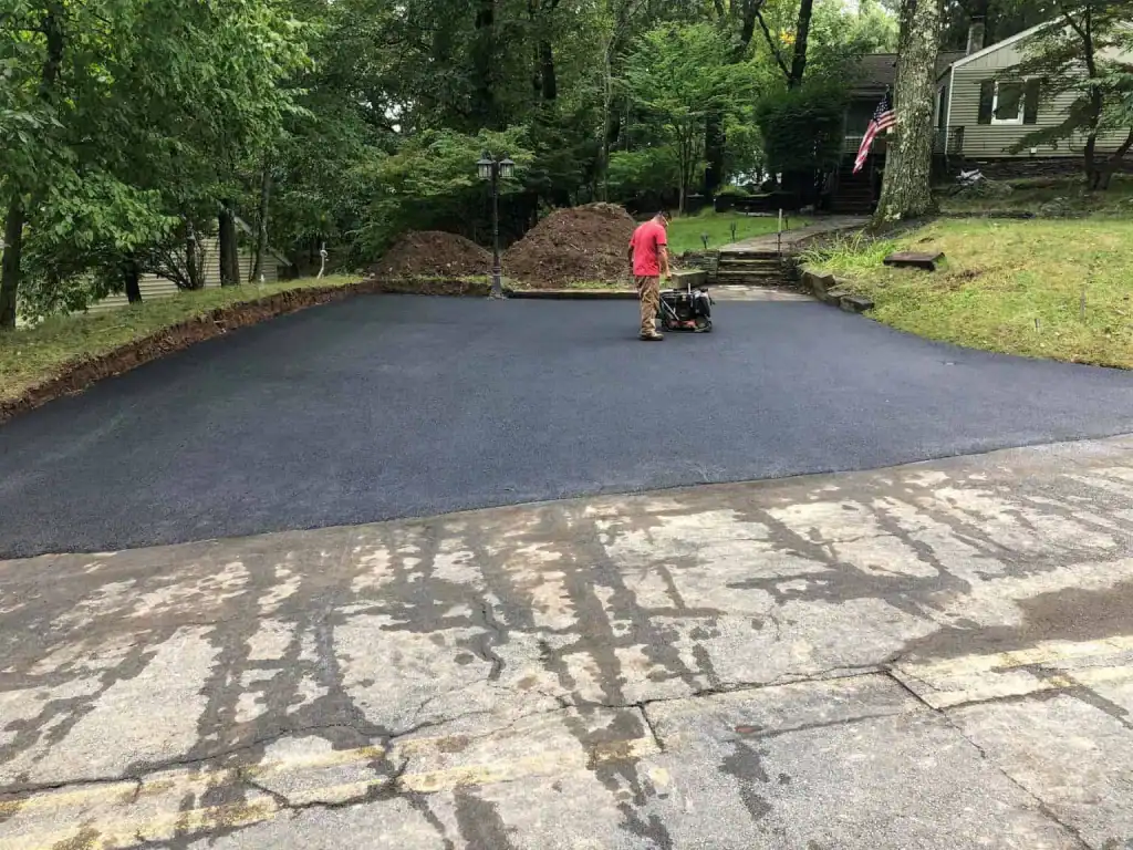 A person in a red shirt operates paving equipment on freshly laid asphalt in a driveway, surrounded by trees and a house with an American flag—showcasing the skilled work of Sussex & Somerset County, NJ paving contractors.