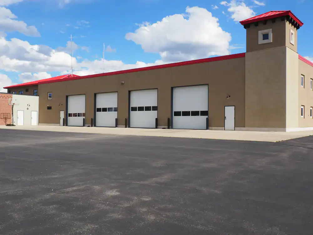 A tan fire station with four large white garage doors, a red roof, and a tall corner tower sits under a blue sky with clouds. The building overlooks an expertly finished asphalt parking lot by paving contractors in Morris, Sussex & Somerset County, NJ.