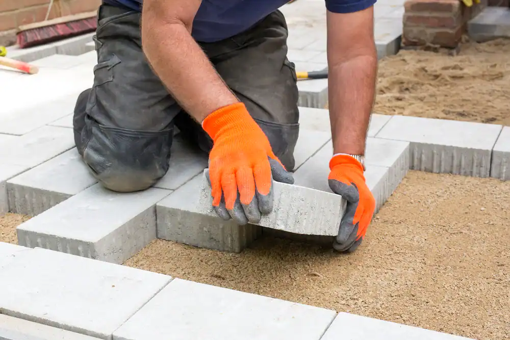 A person wearing orange gloves and dark pants kneels on the ground, placing rectangular concrete pavers onto a bed of sand&mdash;just like expert Paving Contractors in Morris, NJ, Sussex & Somerset County create stunning paved surfaces outdoors.
