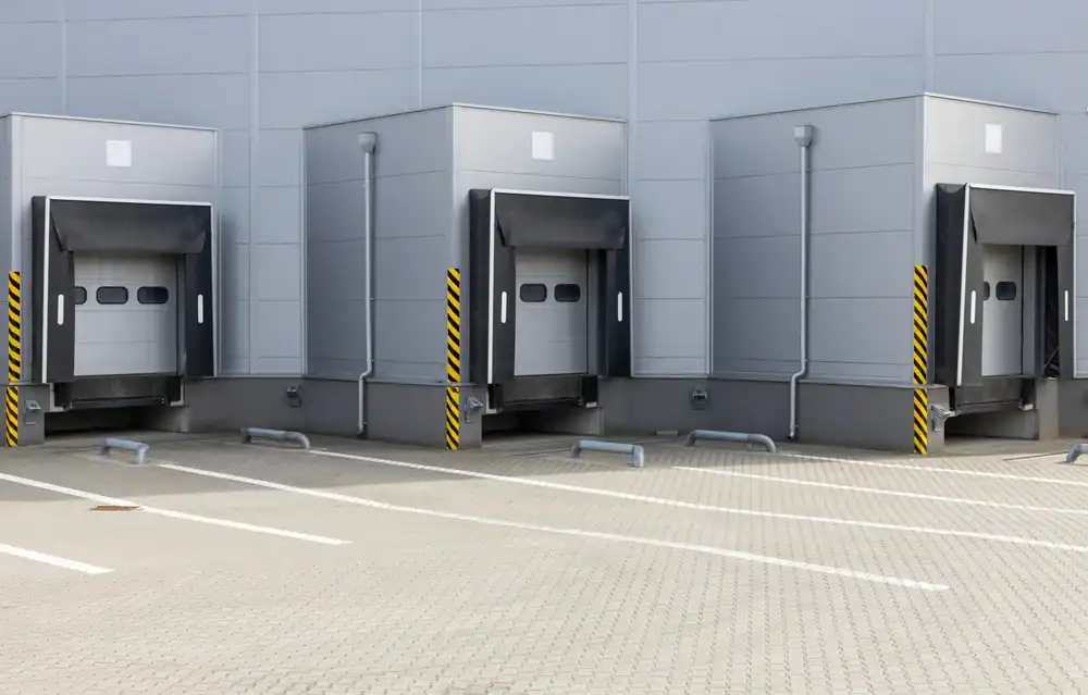 Three empty loading dock bays with closed gray overhead doors, yellow and black striped protective barriers, and marked parking spaces&mdash;expertly finished by paving contractors serving Morris, NJ, Sussex & Somerset County&mdash;set against a light gray industrial building.