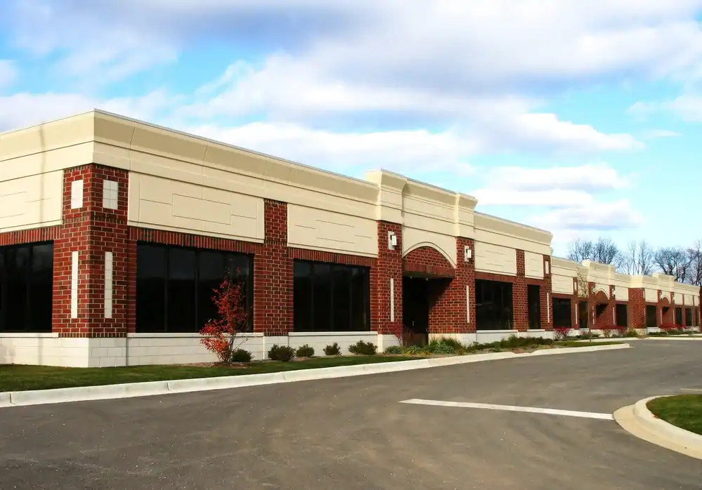 A single-story office building with red brick and beige accents, large dark windows, and a landscaped exterior sits under a partly cloudy sky next to an empty parking lot paved by expert Paving Contractors Morris, NJ.