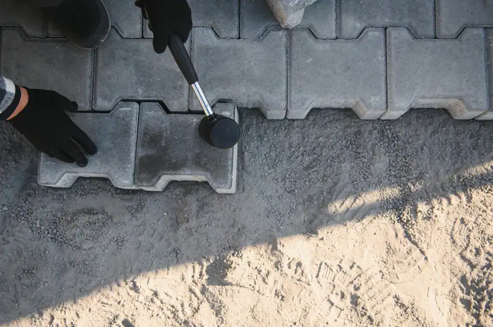 A person in black gloves uses a rubber mallet to install concrete pavers on compacted sand, aligning interlocking blocks for a pathway&mdash;a technique often used by paving contractors in Morris, Sussex & Somerset County, NJ.