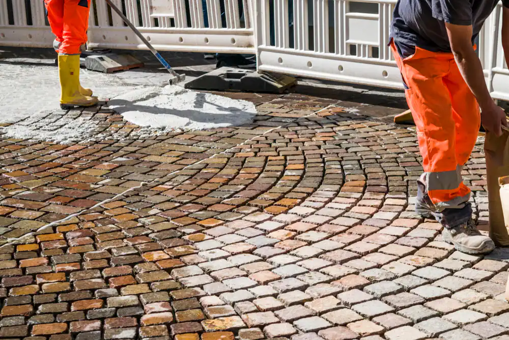 Two workers in bright orange pants repair a cobblestone street in Sussex & Somerset County, NJ. One adjusts stones while the other applies cement or grout, showcasing the skilled craftsmanship of paving contractors Morris. A temporary barrier is visible behind them.