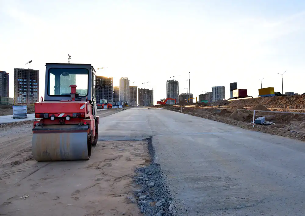 A red road roller sits on a partially paved roadway at a construction site in NJ, with modern high-rise buildings and cranes in the background&mdash;showcasing the work of Paving Contractors Morris, Sussex & Somerset County under a bright sky.