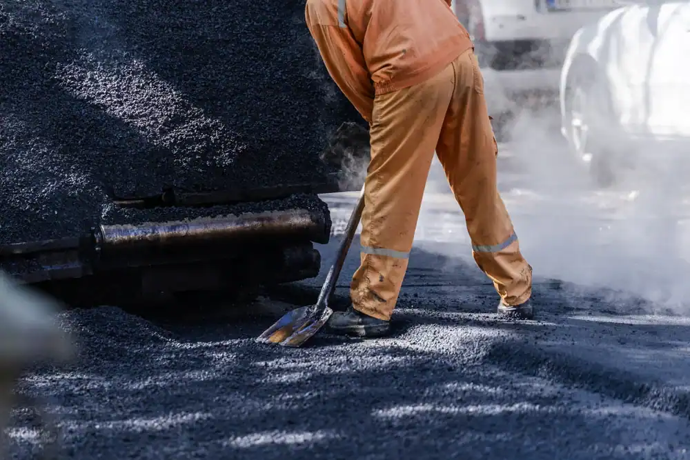 A worker in orange clothing uses a shovel to spread hot asphalt on a road, with steam rising from the surface. Paving Contractors Morris, Sussex & Somerset County, NJ operate nearby machinery and a white vehicle is visible in the background.