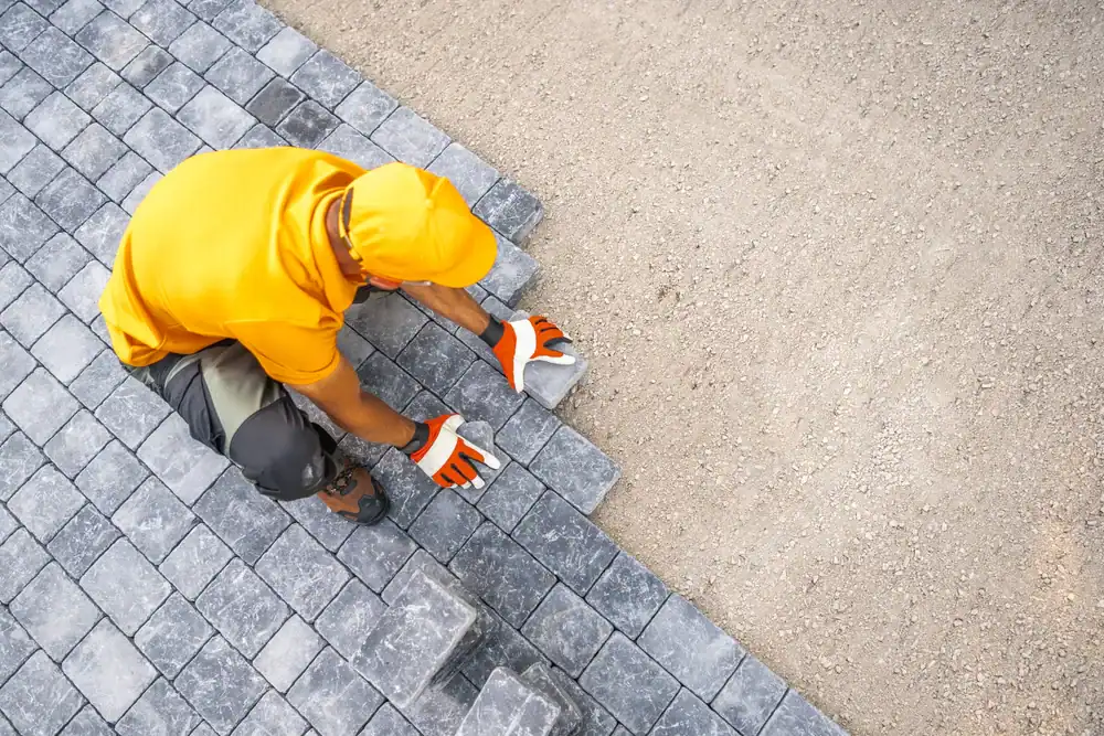 A worker in an orange shirt and gloves is laying gray paving stones on the ground, aligning them next to bare earth. Captured from above, this scene highlights the skilled work of NJ Paving Contractors Morris, Sussex & Somerset County.