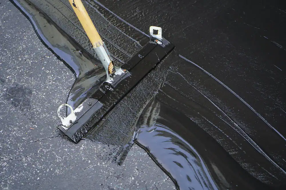 A close-up of a squeegee spreading black sealant over an asphalt driveway, expertly applied by paving contractors in Morris, Sussex & Somerset County, NJ&mdash;part of the surface is freshly coated while the rest remains exposed.